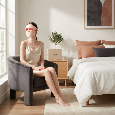 A woman wearing a professional LED red light mask while relaxing in a modern, sunlit bedroom with terracotta and charcoal dec
