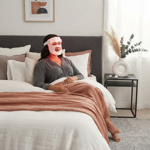 A woman wearing a professional LED red light mask while relaxing on a bed with terracotta and charcoal home decor.