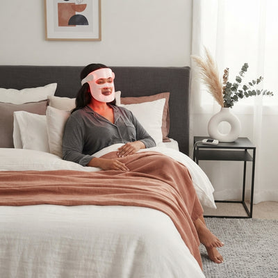 A woman wearing a professional LED red light mask while relaxing on a bed with terracotta and charcoal home decor.